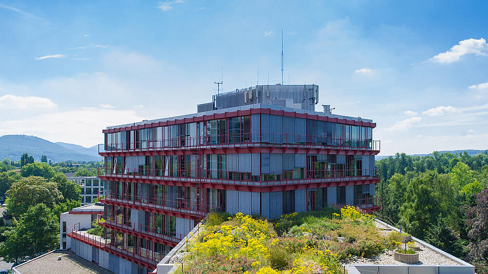 © Frank Homann Außenansicht des Wissenschaftszentrums Bonn. Das moderne Gebäude mit auffälligen roten Fassadenelementen und großen Glasflächen steht inmitten grüner Bäume. Auf dem Dach befindet sich eine begrünte Fläche mit gelb blühenden Pflanzen. Im Hintergrund sind Wälder und Hügel zu erkennen, der Himmel ist blau mit vereinzelten Wolken.