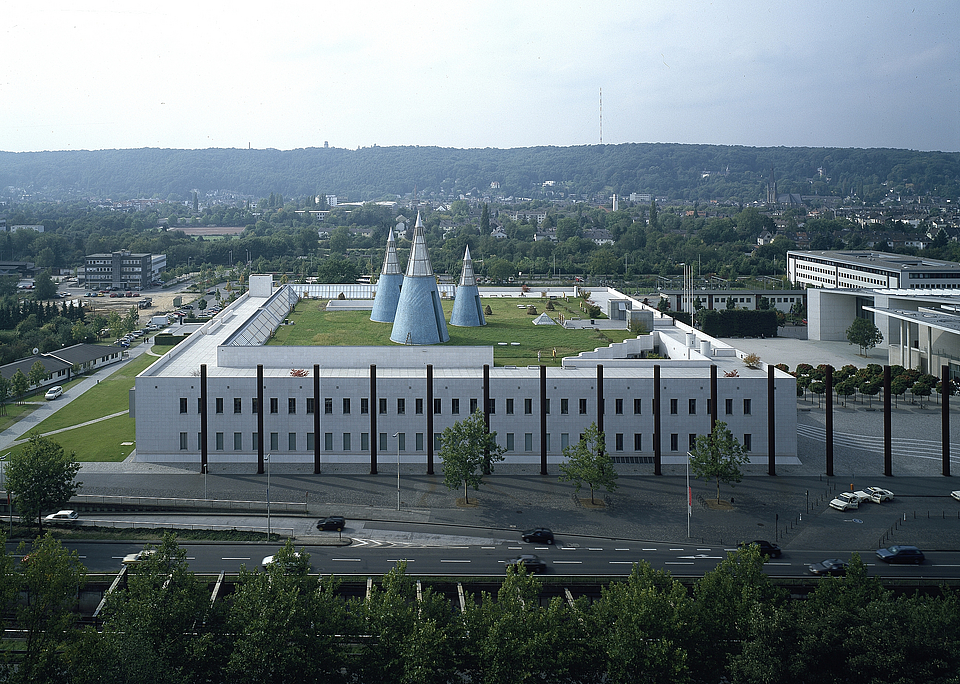 © Bundeskunsthalle Außenansicht der Bundeskunsthalle in Bonn. Das moderne Museumsgebäude hat eine weiße Fassade mit klaren, rechteckigen Strukturen. Auf dem begrünten Dach befinden sich mehrere markante, blau-graue Kegel. Im Vordergrund verläuft eine Straße mit Autos, im Hintergrund ist die Stadt Bonn mit bewaldeten Hügeln zu sehen.